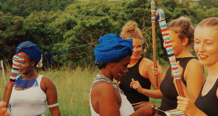 Women laugh and chat while holding brightly beaded sticks during a cultural gathering.