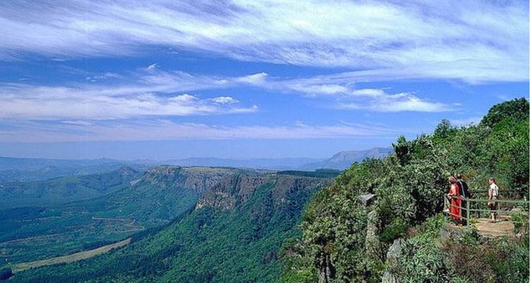 Los visitantes están de pie en un mirador con valla en un acantilado, mirando hacia un amplio valle verde y montañas lejanas.
