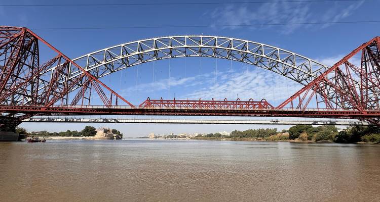 Grand pont en arc d'acier enjambant le large fleuve Indus sous un ciel bleu.