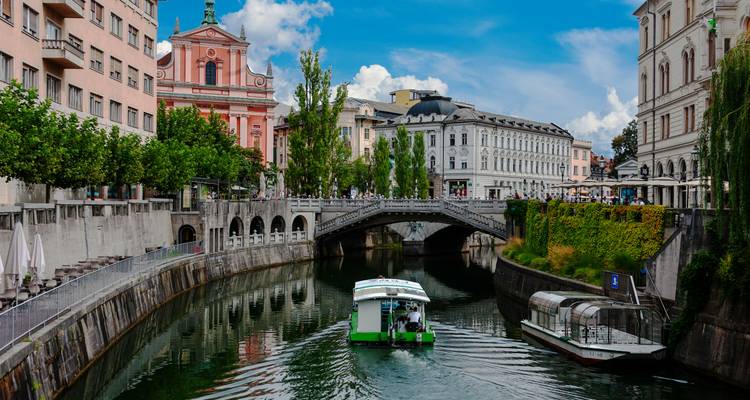Grünes Elektroboot, das unter Ljubljanas Dreifachbrücke mit historischen Fassaden in der Umgebung fährt
