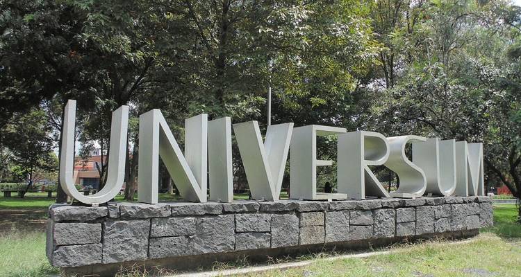 Letrero metálico grande que dice 'UNIVERSUM' colocado en un parque lleno de vegetación sobre una base de piedra.