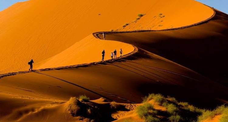 Pequeñas figuras caminan a lo largo de la cresta de una enorme duna de arena naranja en el desierto.