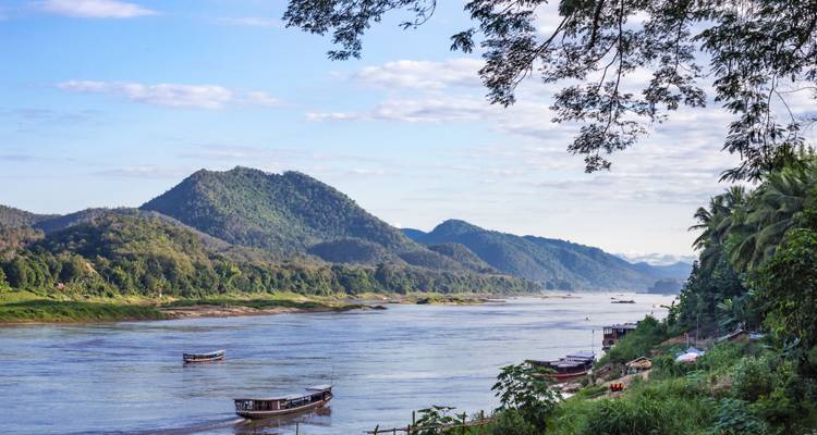 Serene Mekong River scene with longboats drifting past forested mountains under a soft morning sky.