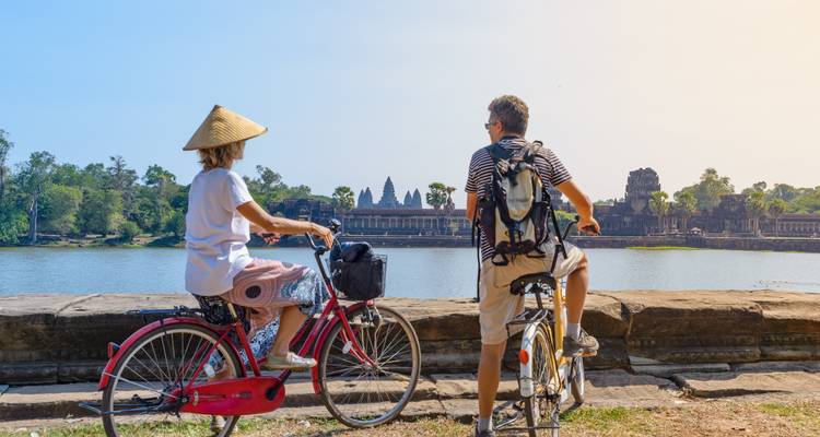 Radfahrer blicken über einen breiten Burggraben hinweg auf die alte Angkor-Anlage im Morgenlicht.