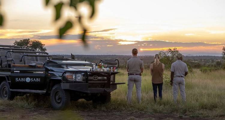 Les rangers et les clients dégustent des boissons au coucher du soleil à côté du véhicule de safari au coucher du soleil sur la brousse.