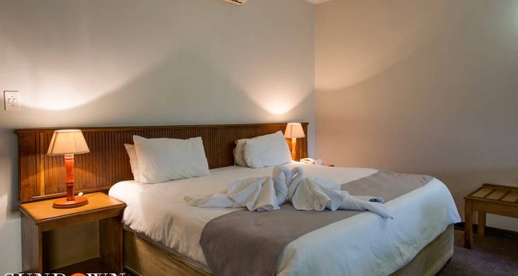 Habitación de hotel simple con un cisne de toalla en la cama e iluminación suave de lámpara; marca de agua tenue de la propiedad en la esquina.