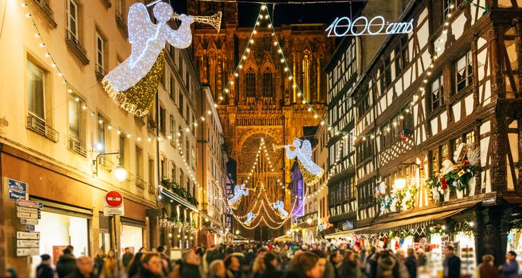 Rue du marché de Noël bondée illuminée par des guirlandes en forme d'anges menant à une cathédrale gothique imposante.