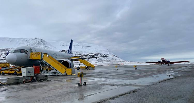 Avión comercial y un avión más pequeño con hélices estacionados en una pista mojada y nevada rodeada de colinas árticas.