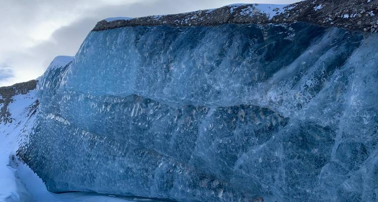 Enorme pared de hielo azul de un glaciar con terreno nevado y accidentado alrededor.