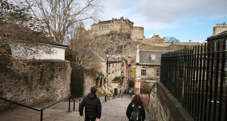 Dos jóvenes viajeros caminan por un callejón empedrado hacia el Castillo de Edimburgo encaramado en la colina.