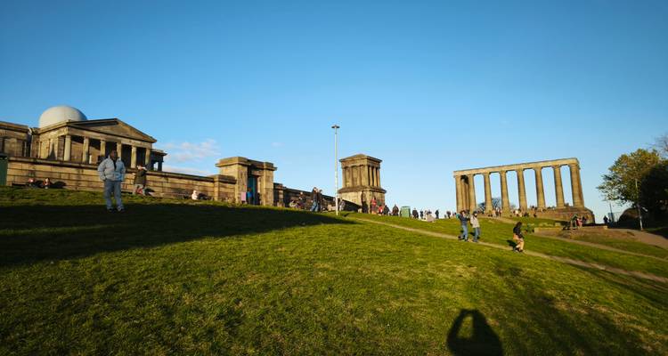 Los visitantes se relajan en las laderas cubiertas de pasto de Calton Hill con el Monumento Nacional y el observatorio bajo un cielo azul despejado.