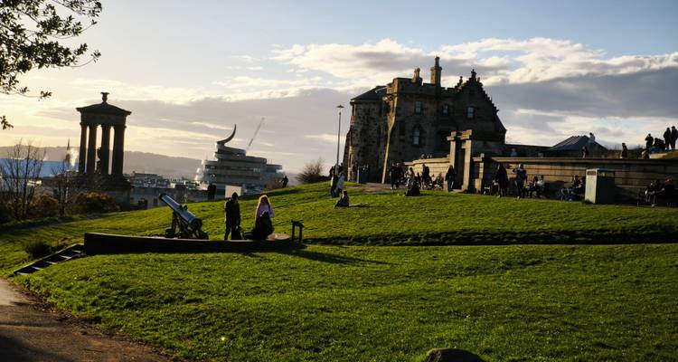 Visitantes recortados contra la luz iluminados por la luz de final de la tarde en Calton Hill con monumentos de fondo.