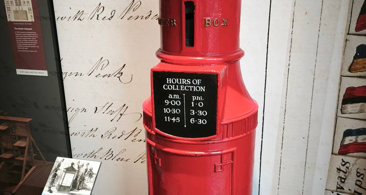 Buzón de correos rojo estilo victoriano en exhibición con placa de horarios de recolección.