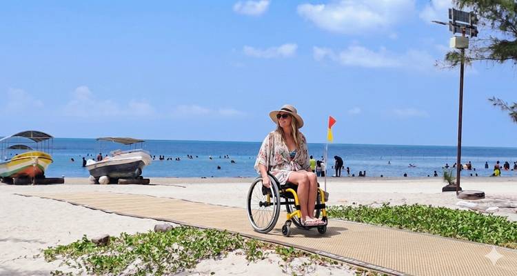 Una mujer en silla de ruedas avanza con confianza por un paseo de madera accesible en una playa tropical de arena con el mar turquesa y el cielo azul.