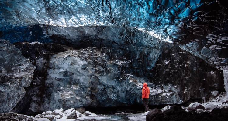Personne vêtue d'une veste rouge se tenant à l'intérieur d'une grotte de glace bleue scintillante aux murs gelés texturés et au sol enneigé.