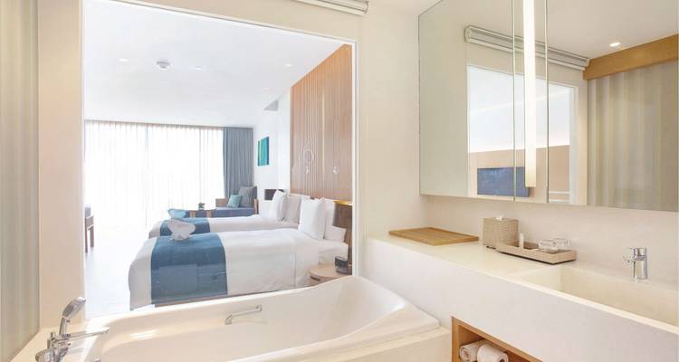 Minimalistisches Badezimmer mit Badewanne und Fenster mit Blick auf ein helles Hotelzimmer mit Doppelbetten.