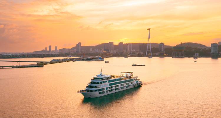 Un crucero blanco se desliza sobre aguas doradas al atardecer con el horizonte de la ciudad de fondo.