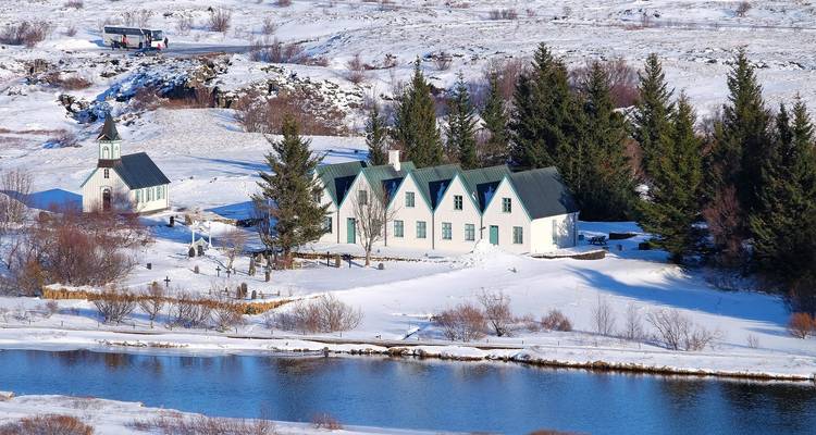 Sneeuwbedekte Thingvellir-kerk en historische huizen naast een reflecterende rivier in een winterlandschap.