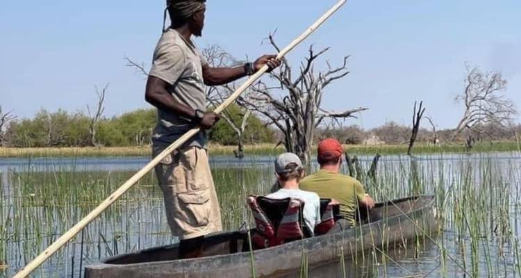 Un guía impulsa una canoa excavada con dos viajeros sentados a través de una vía acuática llena de juncos bajo cielos azules despejados.