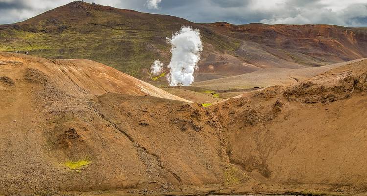 Stoomende geothermische ventilatieopening die uit ruige bruine vulkanische heuvels onder donkere wolken omhoog stijgt.