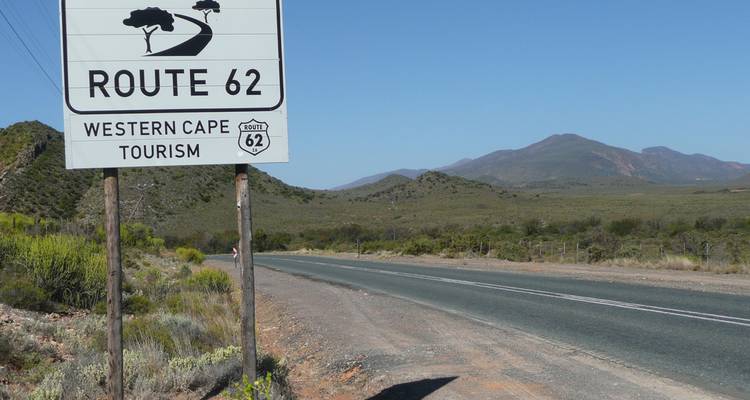 Un letrero indicador de la famosa Ruta 62 en Western Cape se encuentra junto a un camino abierto y colinas distantes.