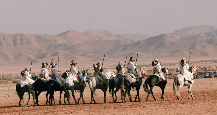 Des cavaliers montés vêtus de robes traditionnelles s'alignent avec de longs fusils sur un fond de montagnes désertiques.