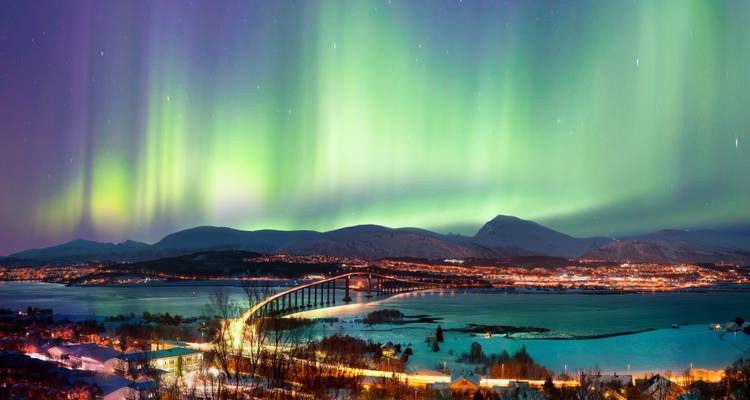 Panorama einer schneebedeckten Küstenstadt, lange Brücke und Fjord, beleuchtet unter lebhaften grünen Nordlichtern.