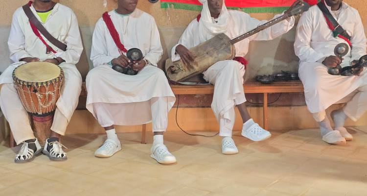 Cuatro músicos en túnicas blancas tocan tambores tradicionales e instrumentos de cuerda dentro de una habitación sencilla.