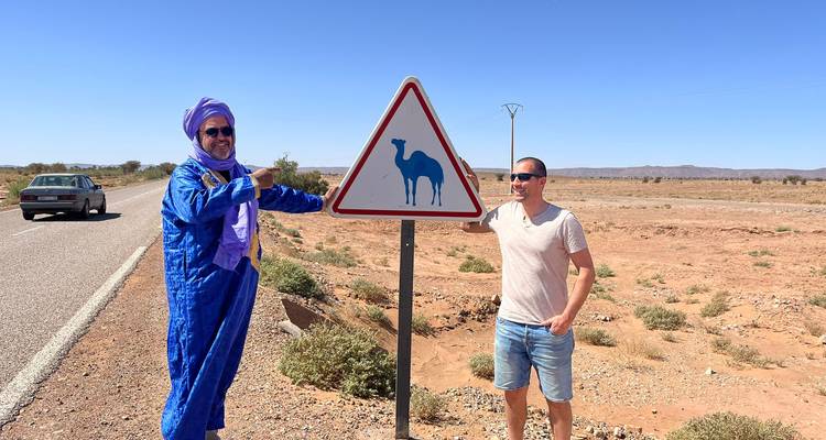Dos viajeros posando junto a una señal triangular de cruce de camellos en una carretera del desierto