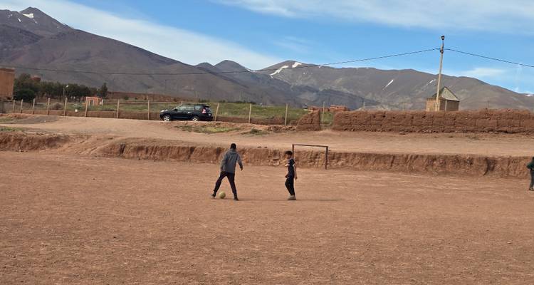 Jóvenes locales jugando fútbol en una cancha de tierra roja con montañas escarpadas de fondo