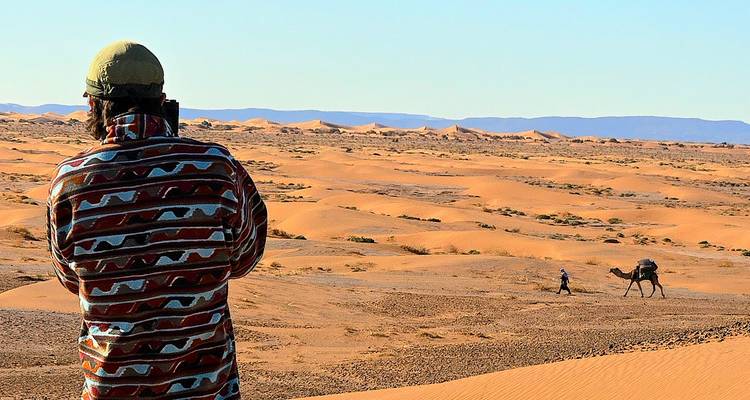 Viajero fotografiando enormes dunas de arena dorada con un camello y su cuidador a lo lejos bajo un cielo azul despejado.