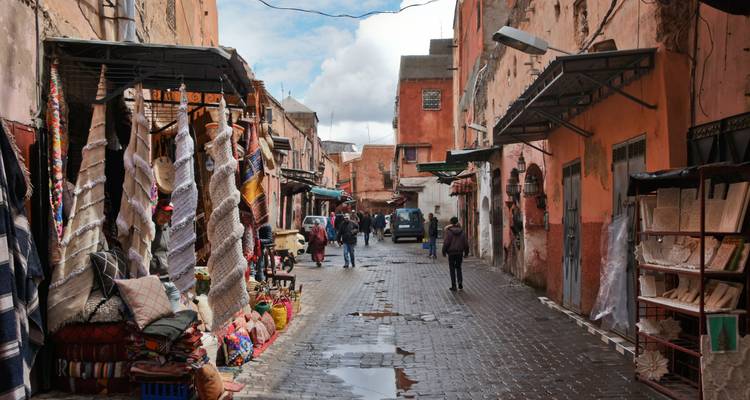 Callejón estrecho y bullicioso del zoco de Marrakech lleno de puestos de textiles y compradores en un día húmedo.