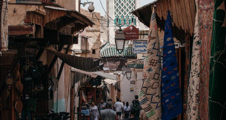 Una estrecha callejuela del zoco en Fez está llena de alfombras colgantes, balcones de madera tallada y visitantes paseando.