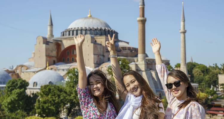 Tres mujeres jóvenes felices posando con los brazos levantados frente a la Basílica de Santa Sofía.