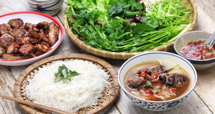 Traditionelle vietnamesische Mahlzeit mit Bun Cha, Nudeln, gegrilltem Schweinefleisch, Kräutern und Dip-Sauce auf einem rustikalen Tisch.