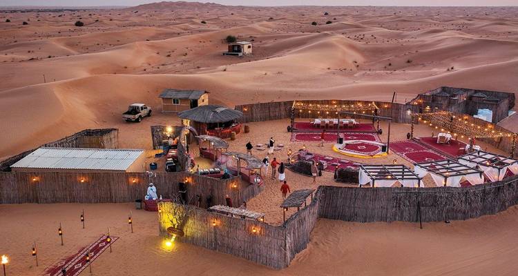Camp bédouin éclairé aux lanternes, entouré de hautes dunes de sable au crépuscule.