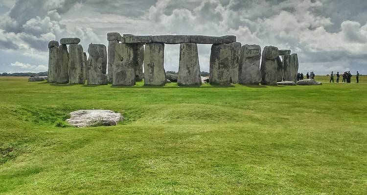 Ikonischer prähistorischer Steinkreis von Stonehenge auf einer weitläufigen Grasfläche unter einem dramatischen bewölkten Himmel, mit einigen entfernten Besuchern zur Größenangabe