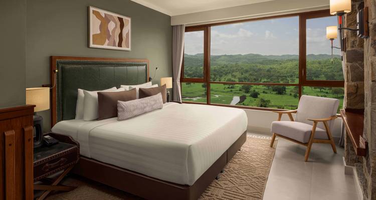 Modern hotel room with king bed and panoramic window overlooking green hills and savannah.