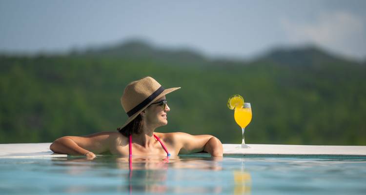 Woman wearing a sun hat relaxes in an infinity pool with a tropical drink against green hills.