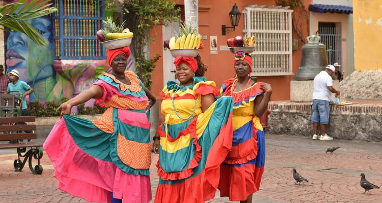 Three brightly dressed Palenquera women pose smiling in a colorful colonial square with murals and pigeons around.