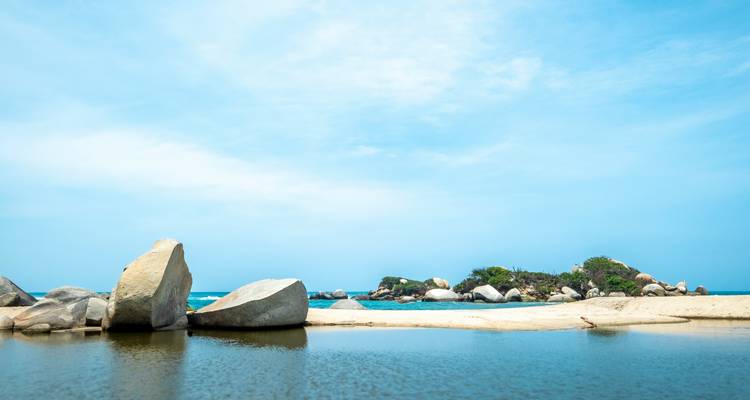 Serene beach lagoon bordered by giant granite boulders and a low sandy spit under a blue sky in Tayrona National Park.