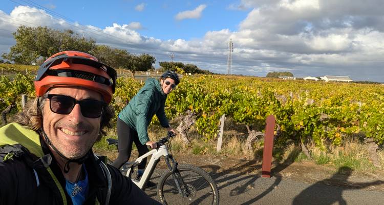 Lachende Reisende halten auf Fahrradern neben herbstlichen Weinbergen unter teilweise bewölktem Himmel an, um ein Selfie zu machen.