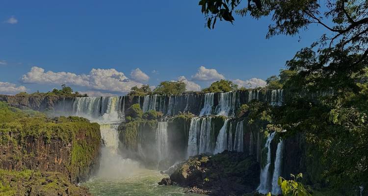 Weitblick auf die Iguazu-Fälle, die über üppige Klippen hinabstürzen und von Regenwald umgeben sind, unter klarem blauen Himmel