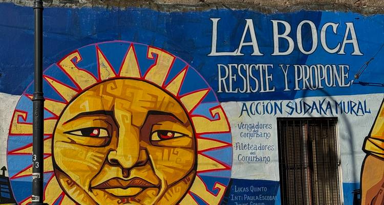 Helles Wandgemälde in La Boca mit der Aufschrift "La Boca Resiste y Propone", stilisiertem Sonnengesicht und blauem Hintergrund