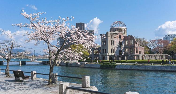 Kirschblüten rahmen die Hiroshima-Friedensdenkmal-Kuppel neben einem ruhigen Fluss unter blauem Himmel ein.