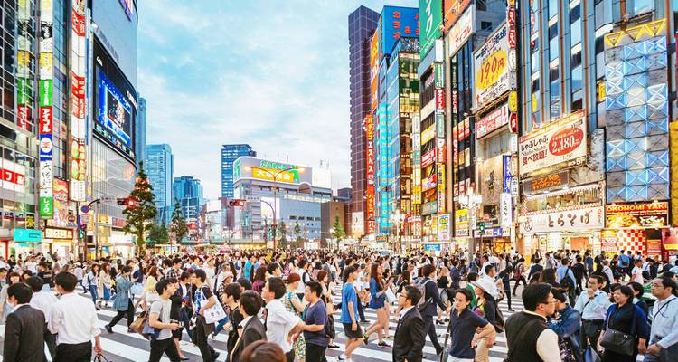 Menschenmengen von Fußgängern, die eine belebte, neonbeleuchtete Kreuzung überqueren, umgeben von Hochhäusern in einer modernen japanischen Stadt.
