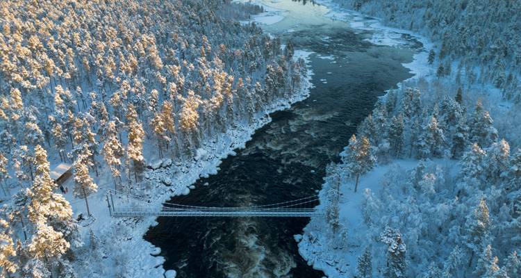 Luchtfoto uit winter van een hangebrug die een gedeeltelijk bevroren rivier overspant, omgeven door sneeuwbedekte pijnbomen.