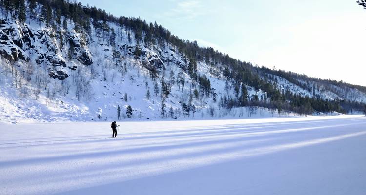 Een skiër glijdt over een uitgestrekt bevroren meer onder steile, sneeuwbedekte rotsen en dennenbossen.