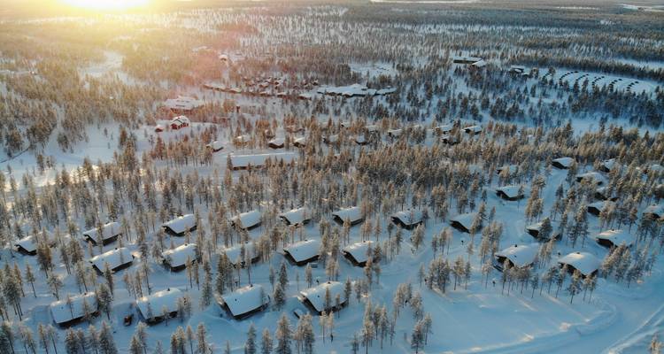 Dronefoto van een sneeuwwoud dorpje met verspreide huisjes onder warm zonnlicht bij zonsopgang.