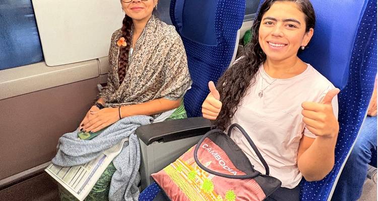 Twee vrouwen zittend in een trein die een duim omhoog geven, gekleed in casual reiskleding en met tassen in hun handen.